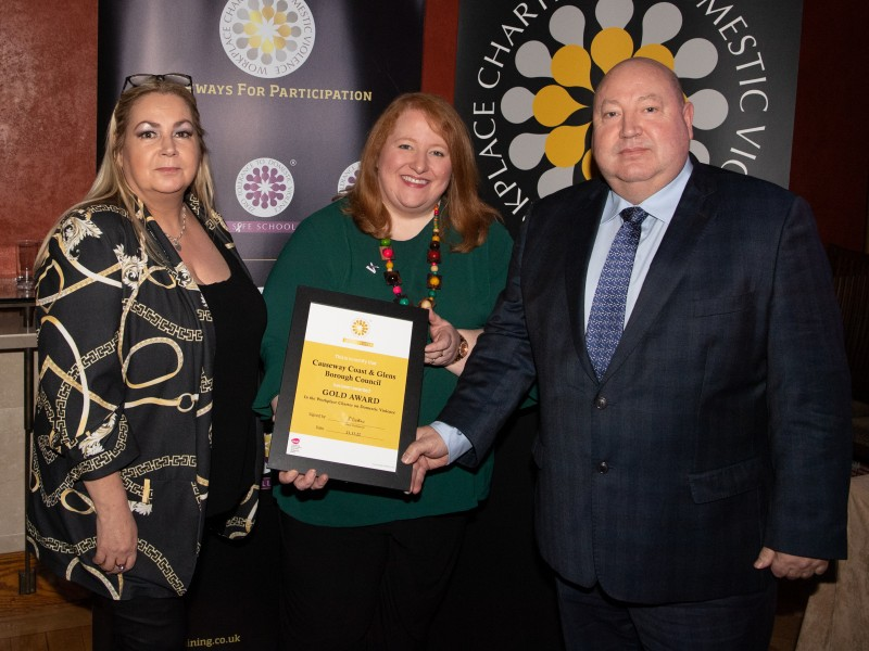 The Chair of Causeway Coast and Glens Policing and Community Safety Partnership, Alderman Adrian McQuillan, receives the Onus Gold Workplace Charter Award from Naomi Long, along with PCSP member Patricia McQuillan.