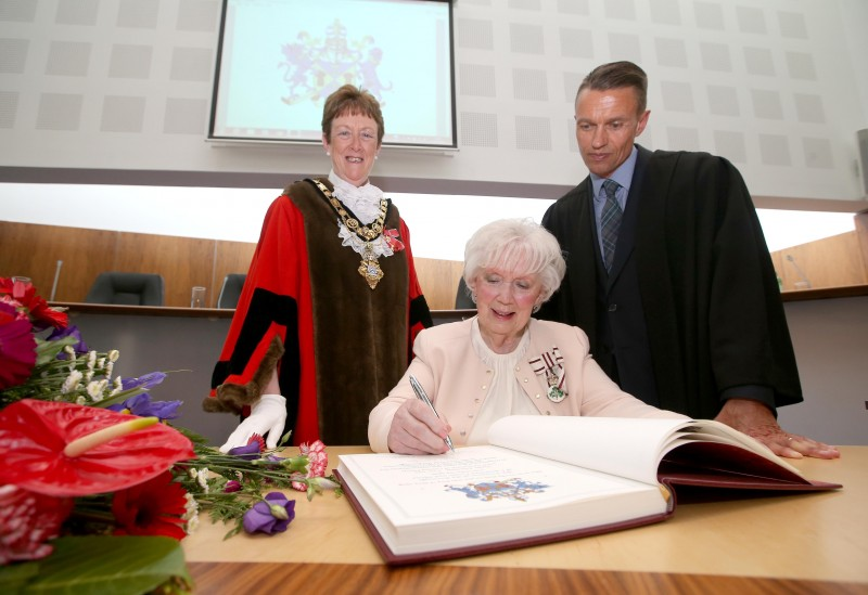 Mrs Joan Christie CVO OBE signs the Freedom register at her Freedom of the Borough ceremony alongside the Mayor of Causeway Coast and Glens Borough Council Councillor Joan Baird OBE and Council Chief Executive David Jackson.